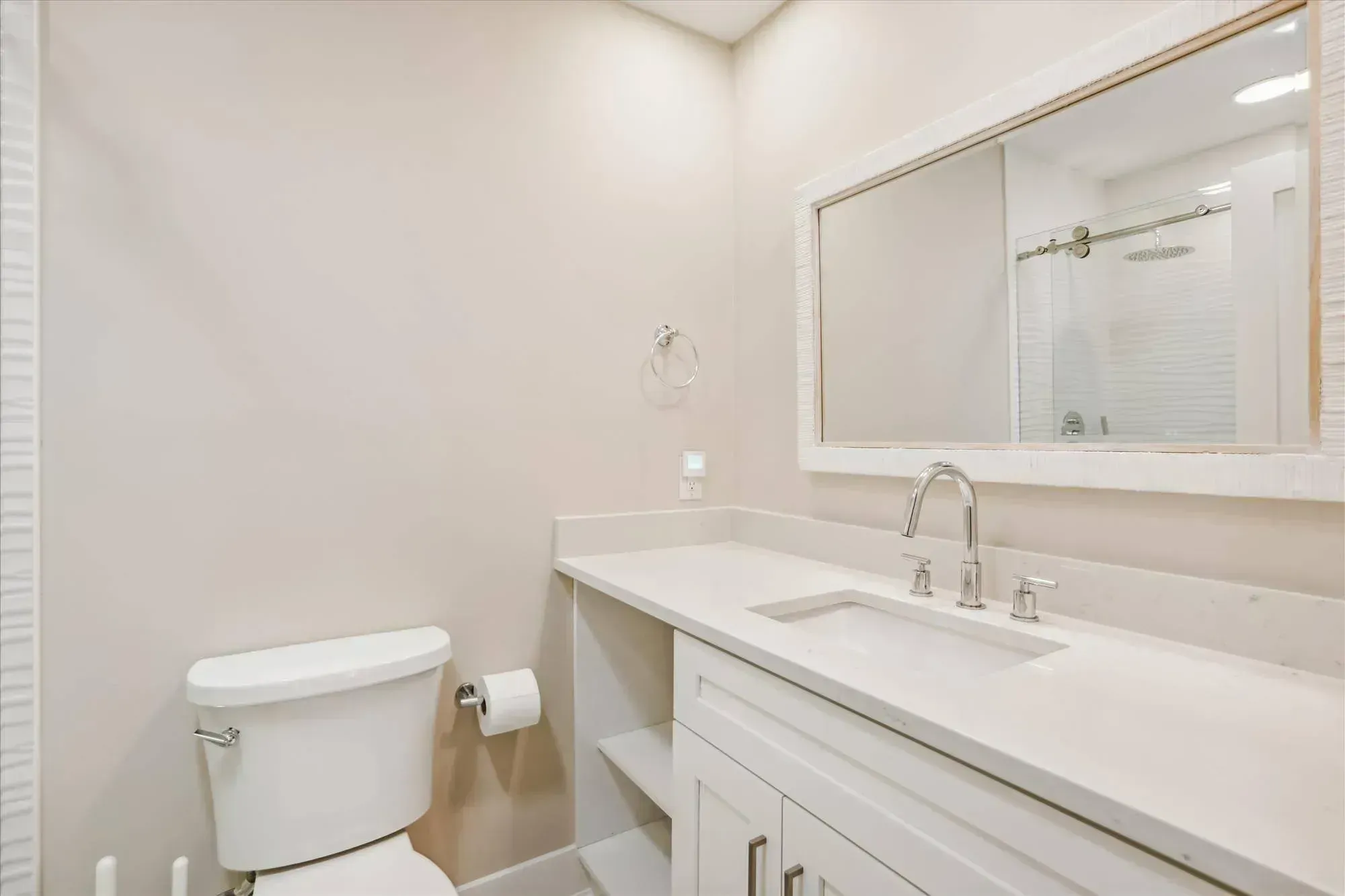 A remodeled bathroom features a white quartz vanity with a chrome faucet and undermount sink beneath a large mirror, with a toilet and glass-enclosed shower reflected nearby.