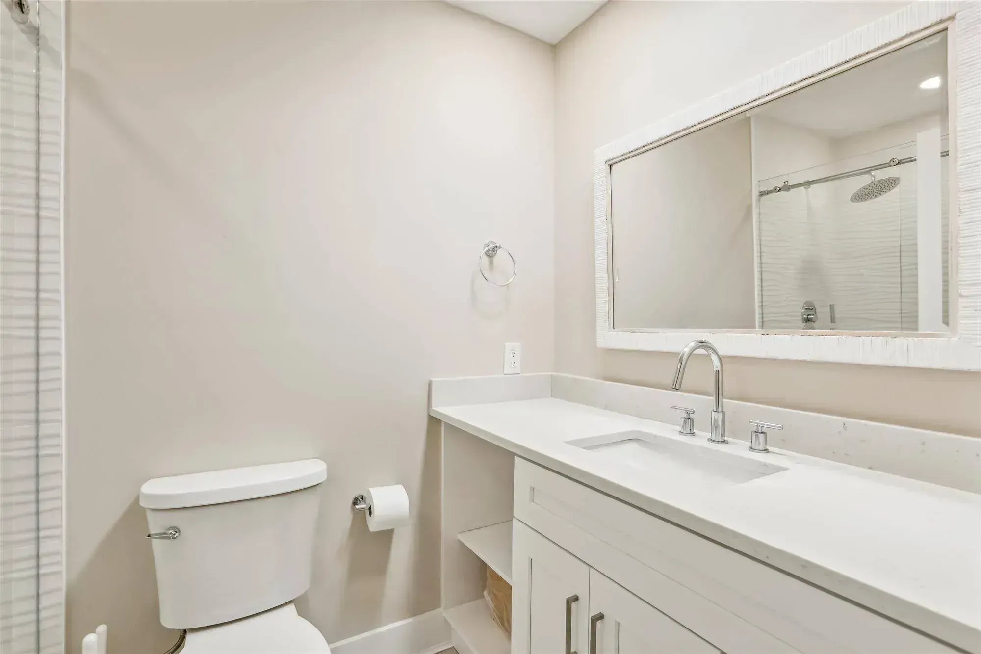 A remodeled bathroom features a white shaker-style vanity with a quartz countertop, a large framed mirror, a toilet, and a glass-enclosed shower with a rain showerhead.