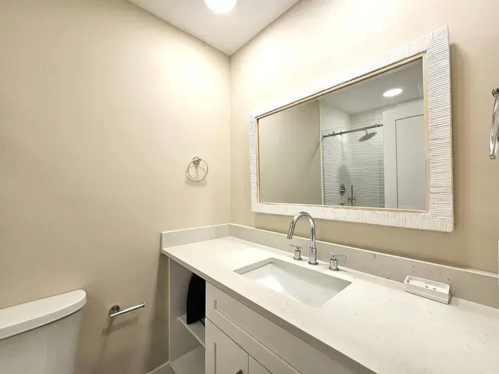 A remodeled bathroom features a white quartz vanity with an undermount sink, chrome fixtures, and a large mirror reflecting a glass-enclosed shower with textured tile.