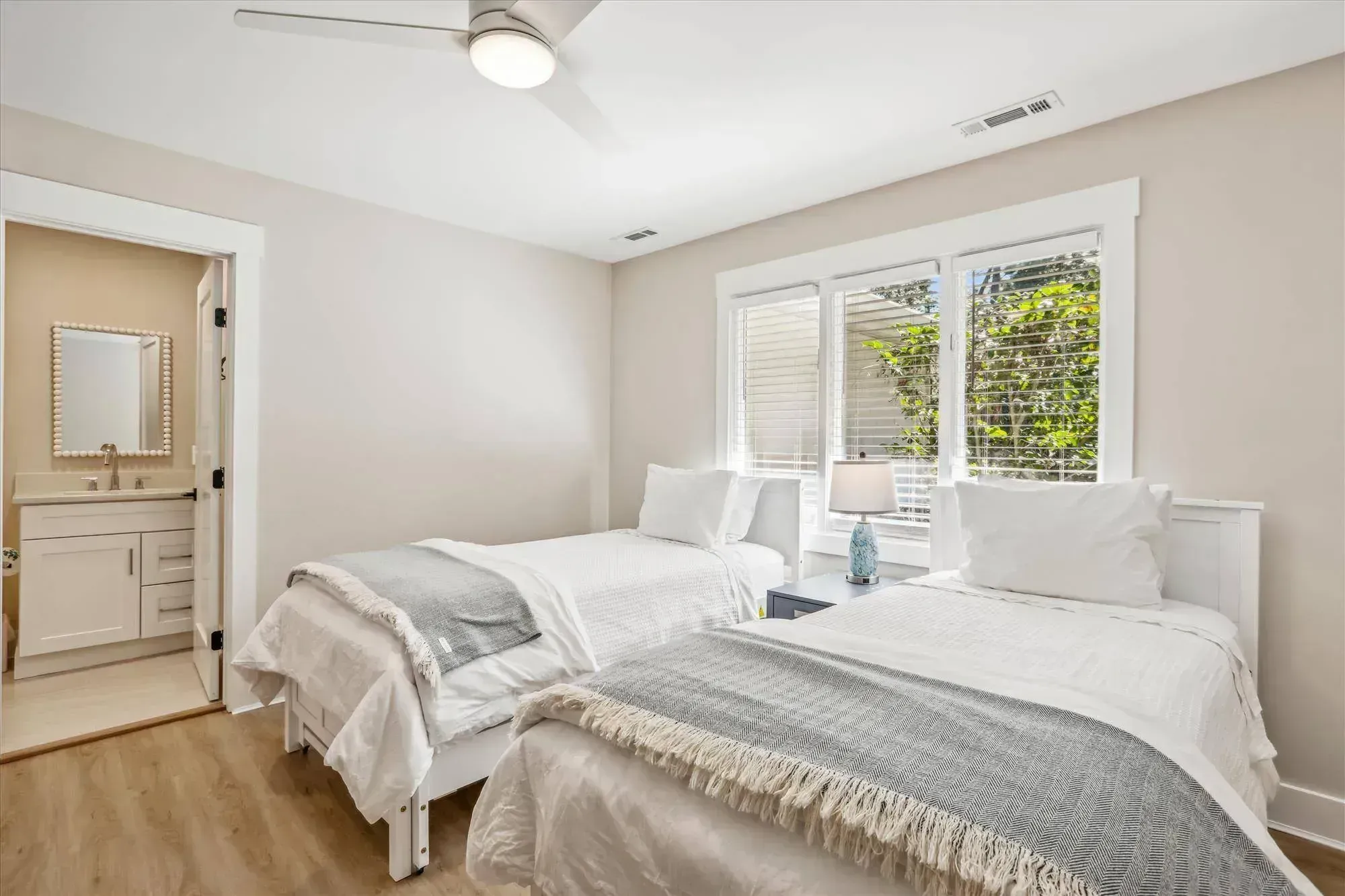 A bright guest bedroom features two twin beds with white and gray bedding, a wood-look floor, large windows with blinds, and a doorway leading to an attached bathroom with a white vanity.
