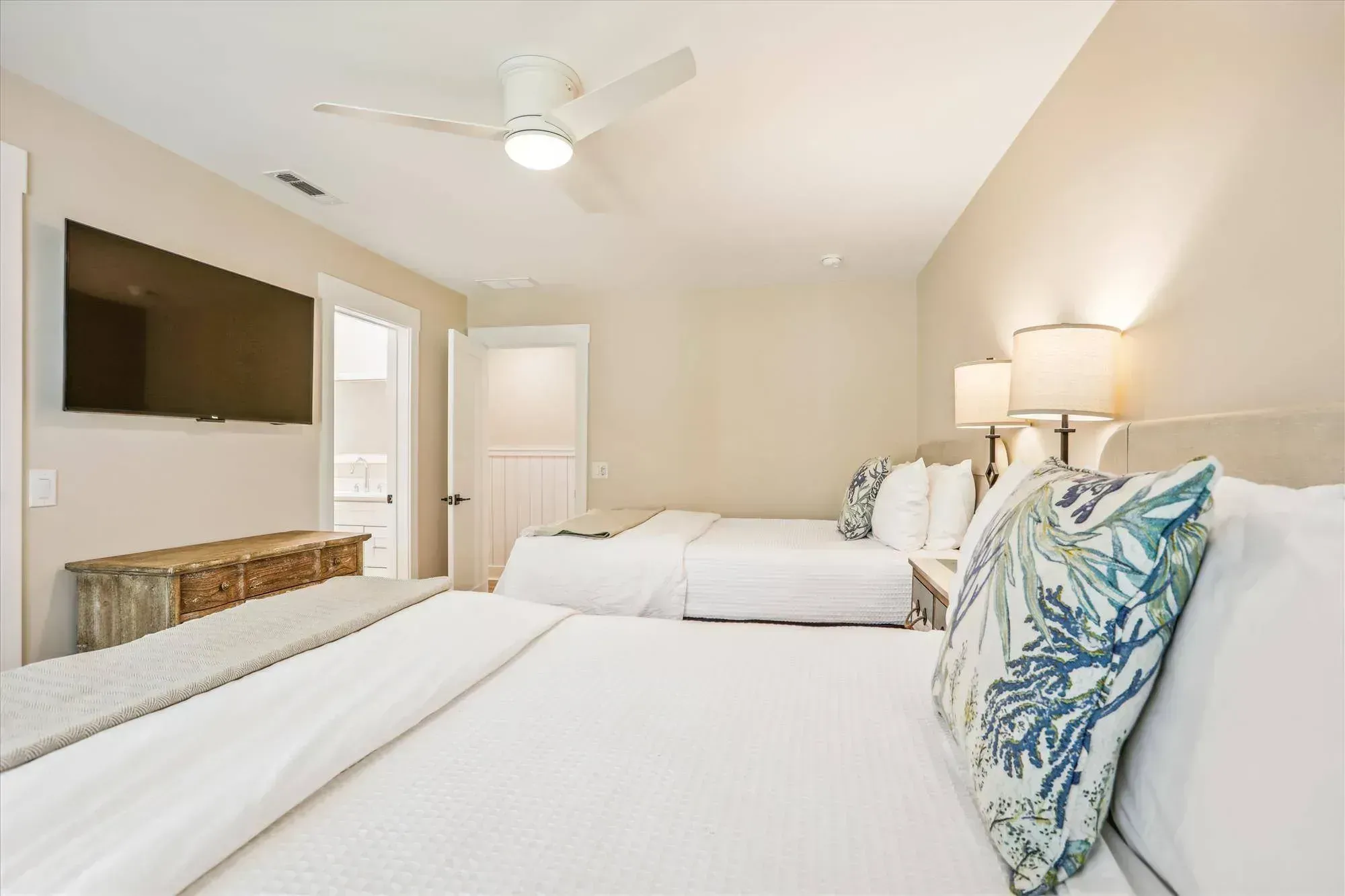 A bright guest bedroom features two beds with white bedding and coastal-patterned pillows, a wall-mounted television, a wooden dresser, and an en-suite bathroom visible through an open door.