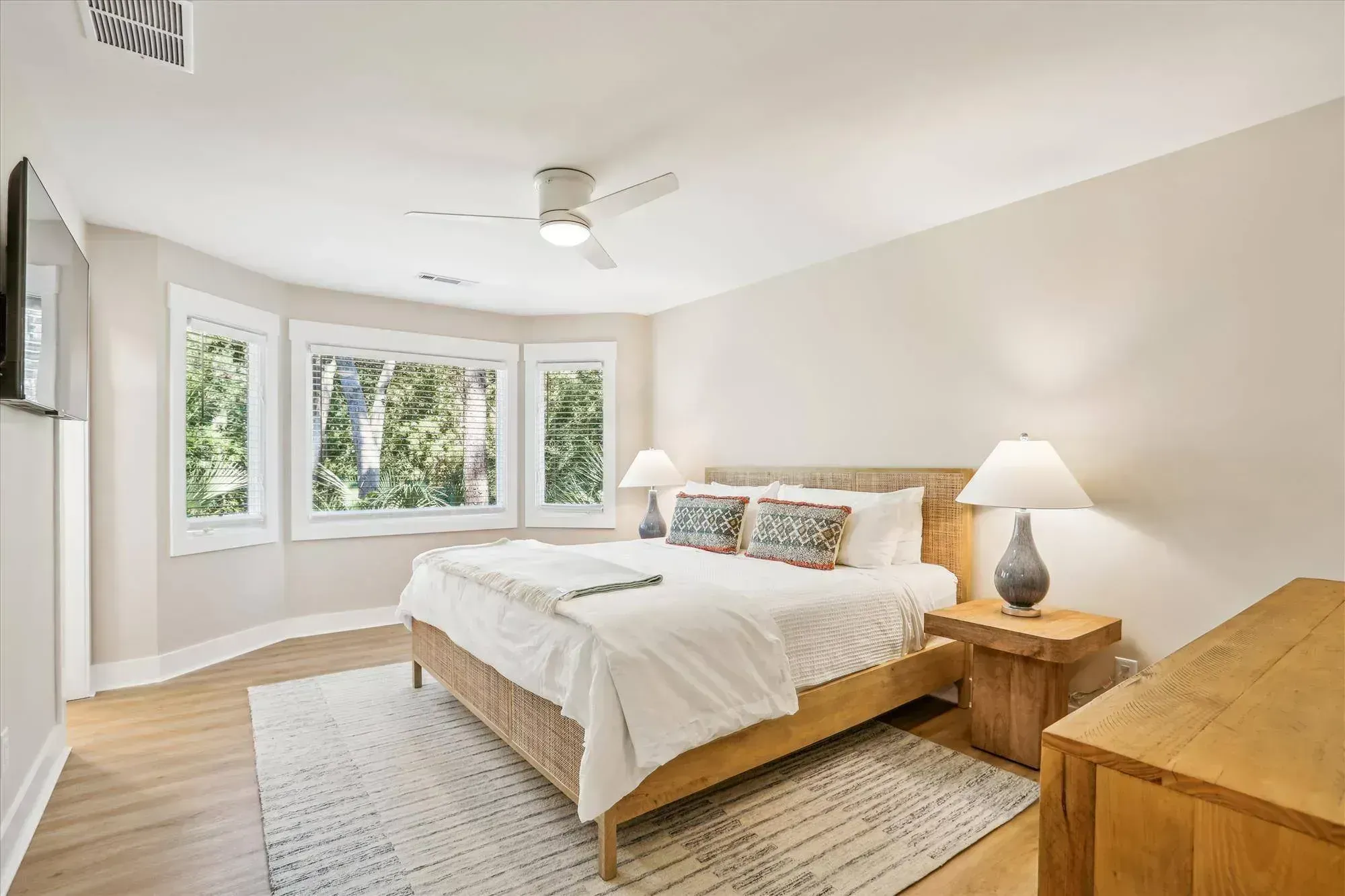 A bright bedroom features a woven wood bed frame with white linens, flanked by two ceramic lamps, in front of a bay window overlooking wooded greenery.