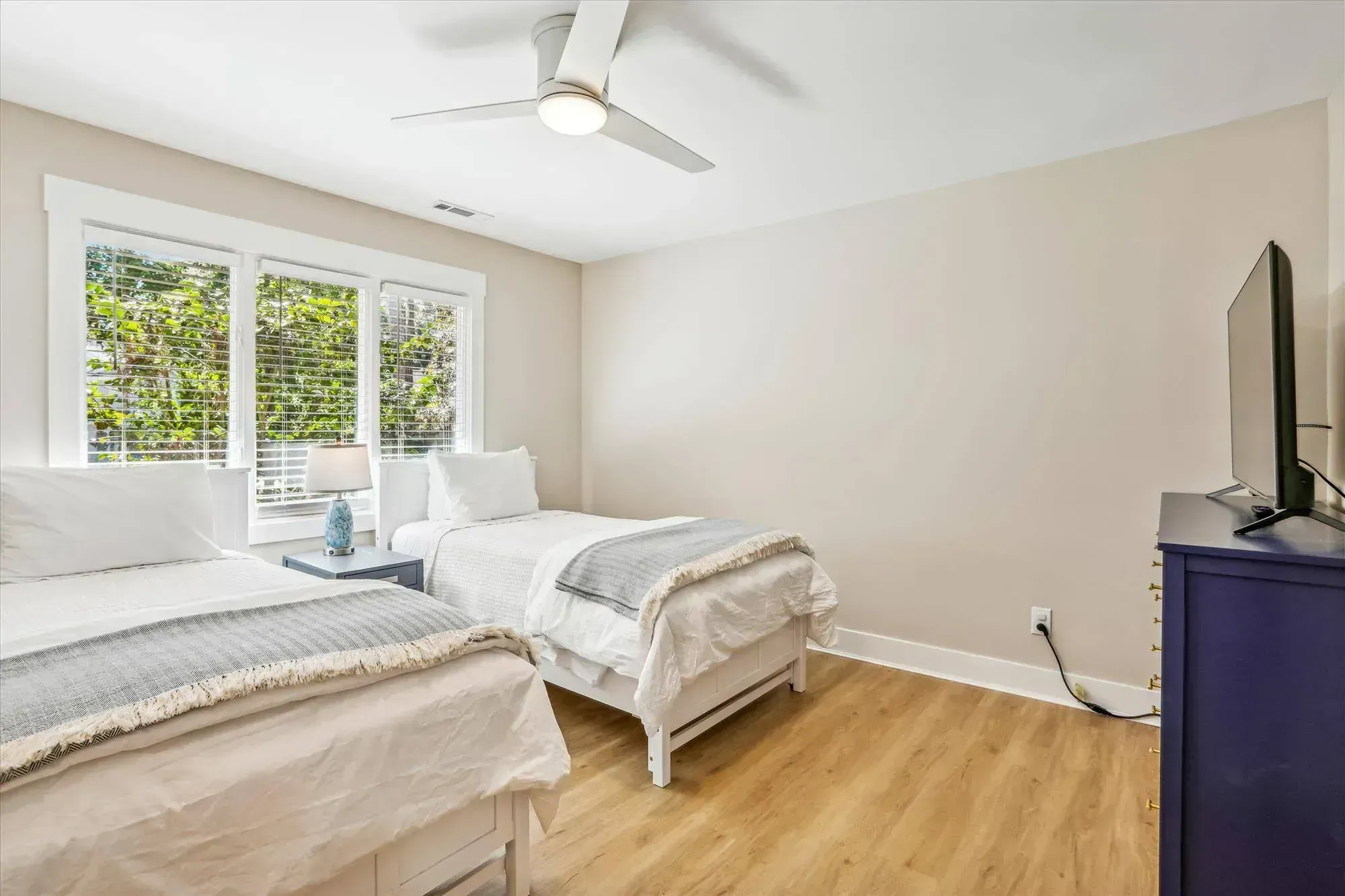 A guest bedroom with two twin beds, a blue nightstand and dresser, wood-look flooring, and a large window overlooking greenery.
