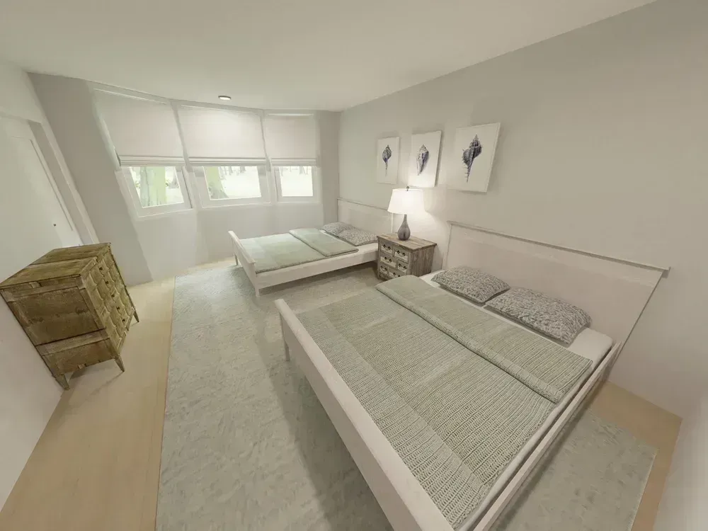 A coastal-themed guest bedroom features two beds with sage green bedding, a bank of windows with shades, a wooden dresser, and framed seashell art on the wall.