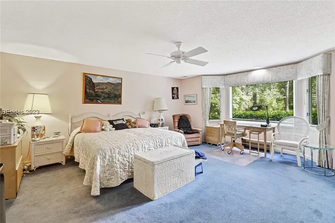 A carpeted bedroom with a large bed, ceiling fan, and a bay window seating area overlooking green trees outside.