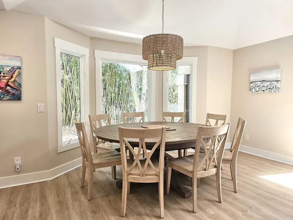 A round wooden dining table surrounded by eight light wood chairs sits beneath a beaded drum chandelier in a bright dining room with a bay window overlooking bamboo landscaping.