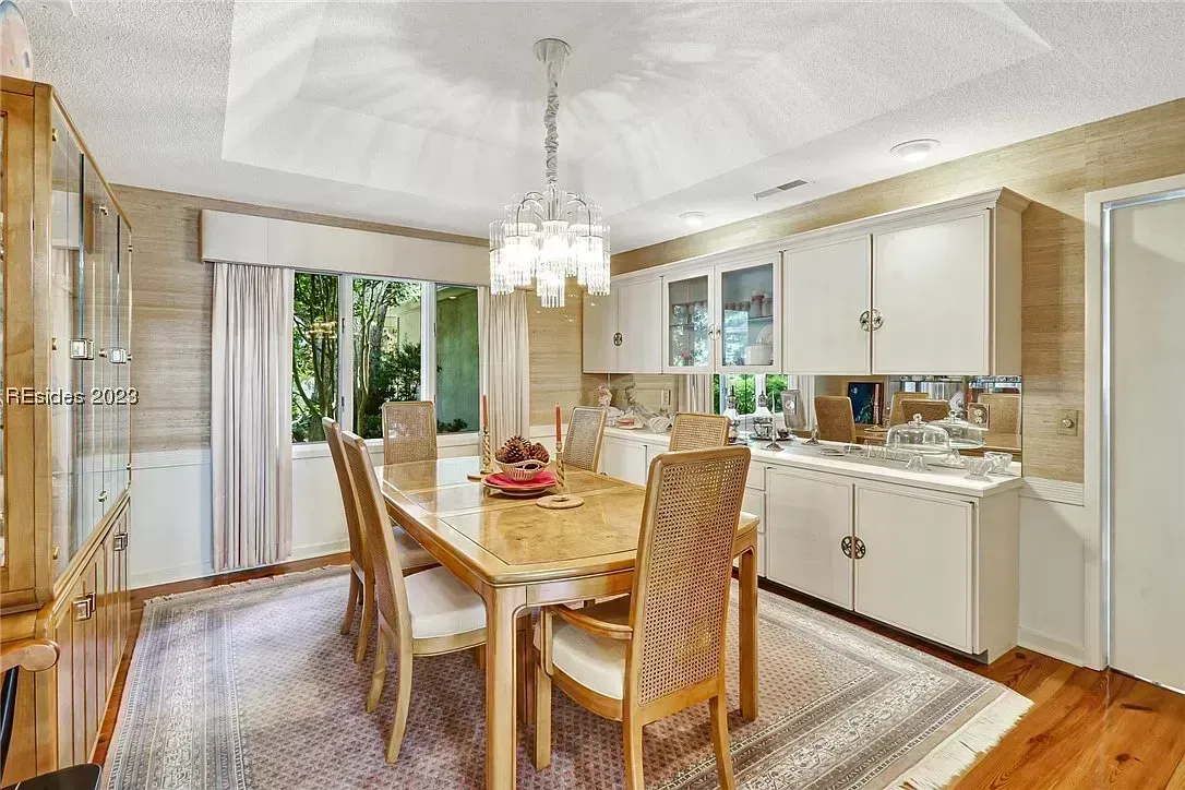 A dining room with a wood table and cane-back chairs sits on a patterned area rug beneath a crystal chandelier, with white built-in cabinetry and a mirrored backsplash along the wall.