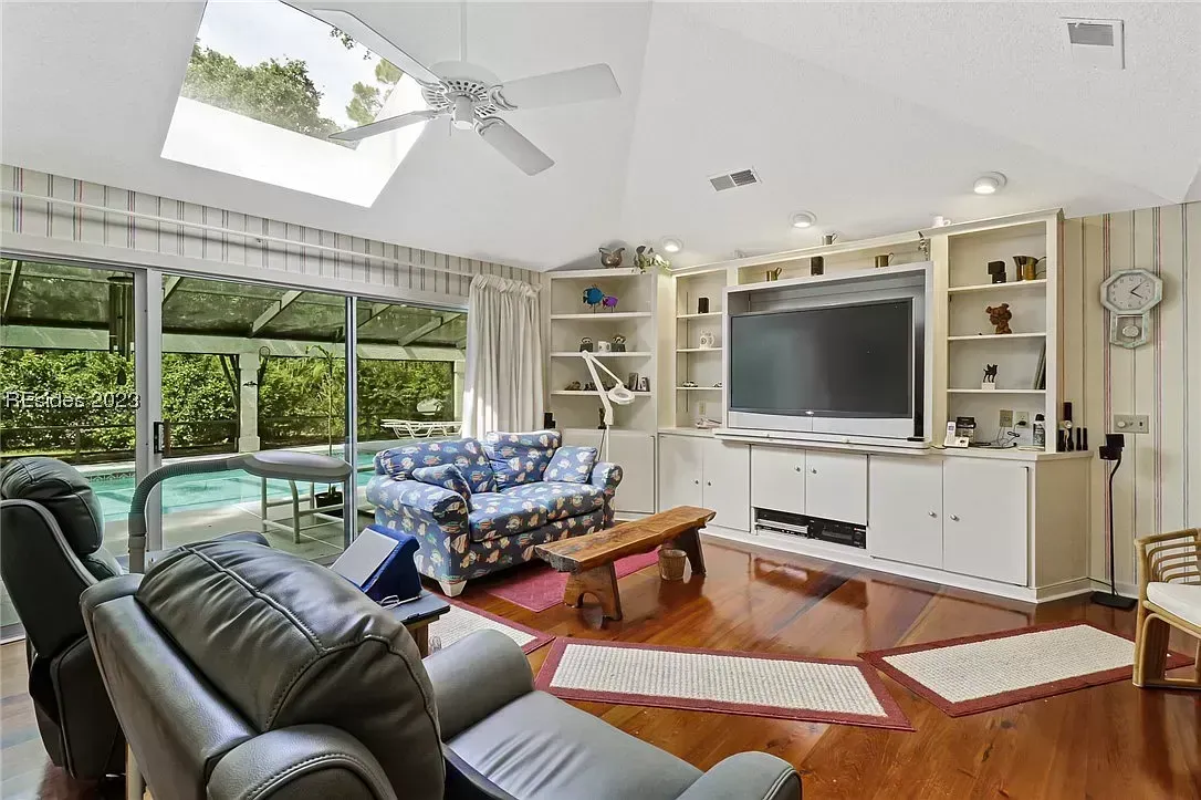 A vaulted family room with a skylight and ceiling fan features a built-in white entertainment center, wood floors, and sliding glass doors that open to a screened pool area.