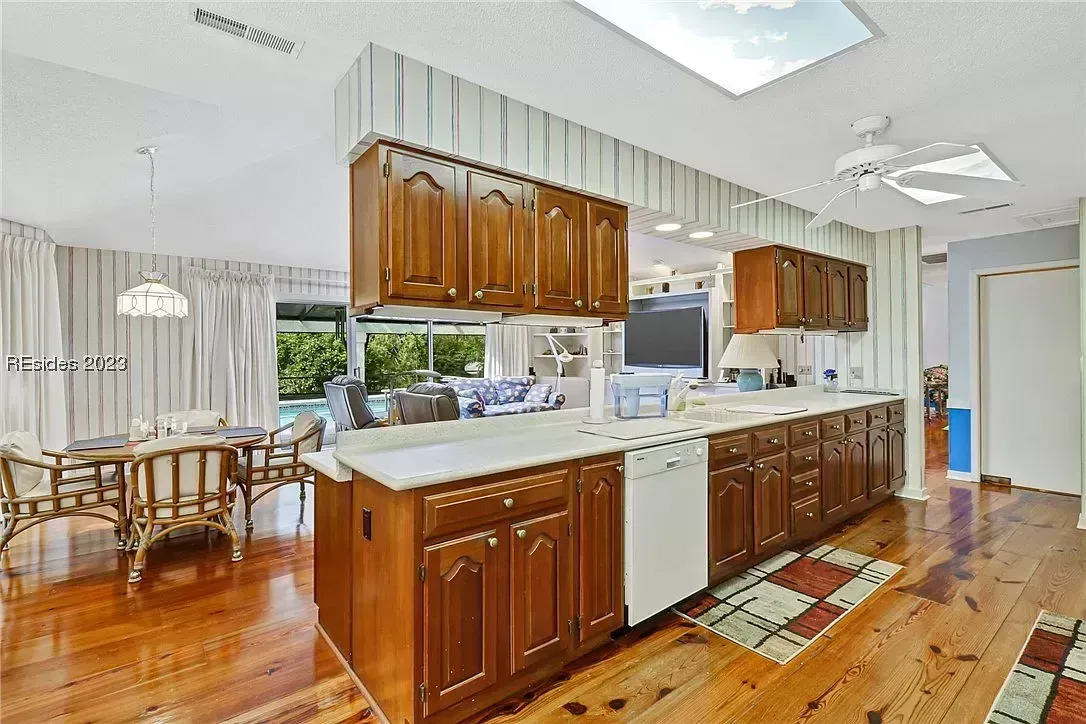 An outdated kitchen with dark wood cabinetry and a white peninsula counter opens into a sitting area with a view of a pool through sliding glass doors, all set on hardwood flooring.