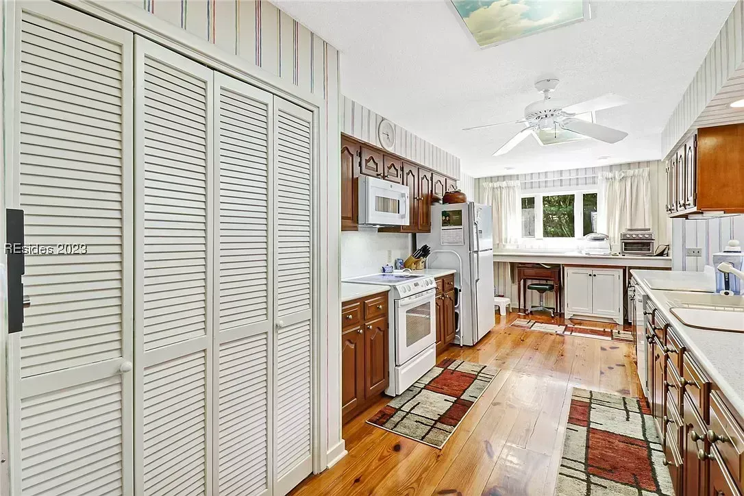 An existing galley kitchen features dark wood cabinetry, white louvered closet doors, white appliances, patterned wallpaper, and a hardwood floor with layered area rugs leading to a window overlooking trees.