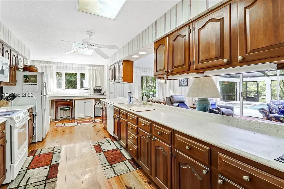 An outdated galley kitchen with dark wood cabinetry and light countertops opens onto a sunlit living area and screened pool beyond sliding glass doors.