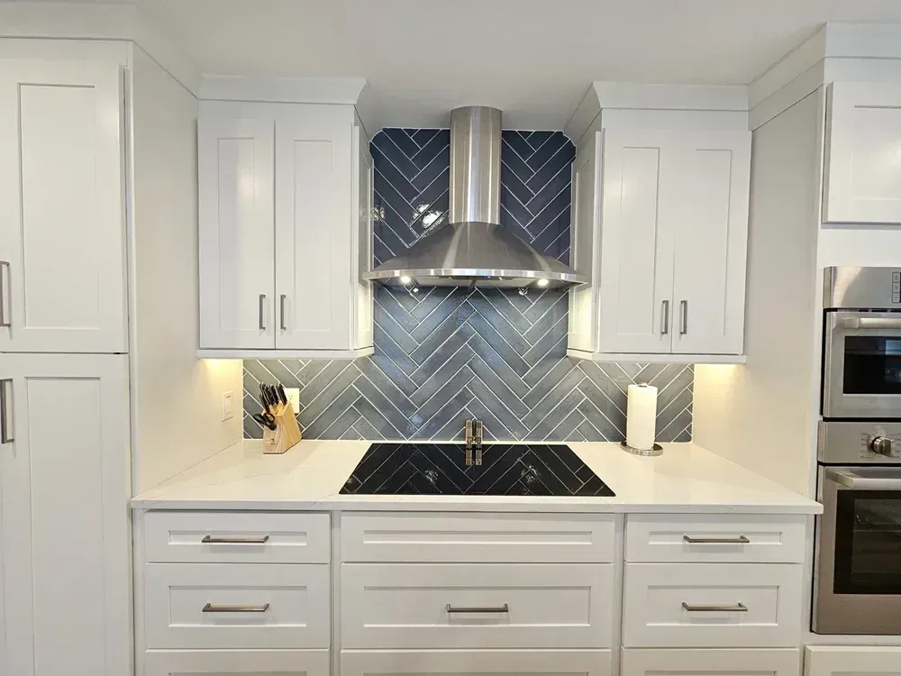 A white shaker cabinet kitchen features a gray herringbone tile backsplash behind the cooktop with a stainless steel chimney hood and built-in wall ovens to the side.