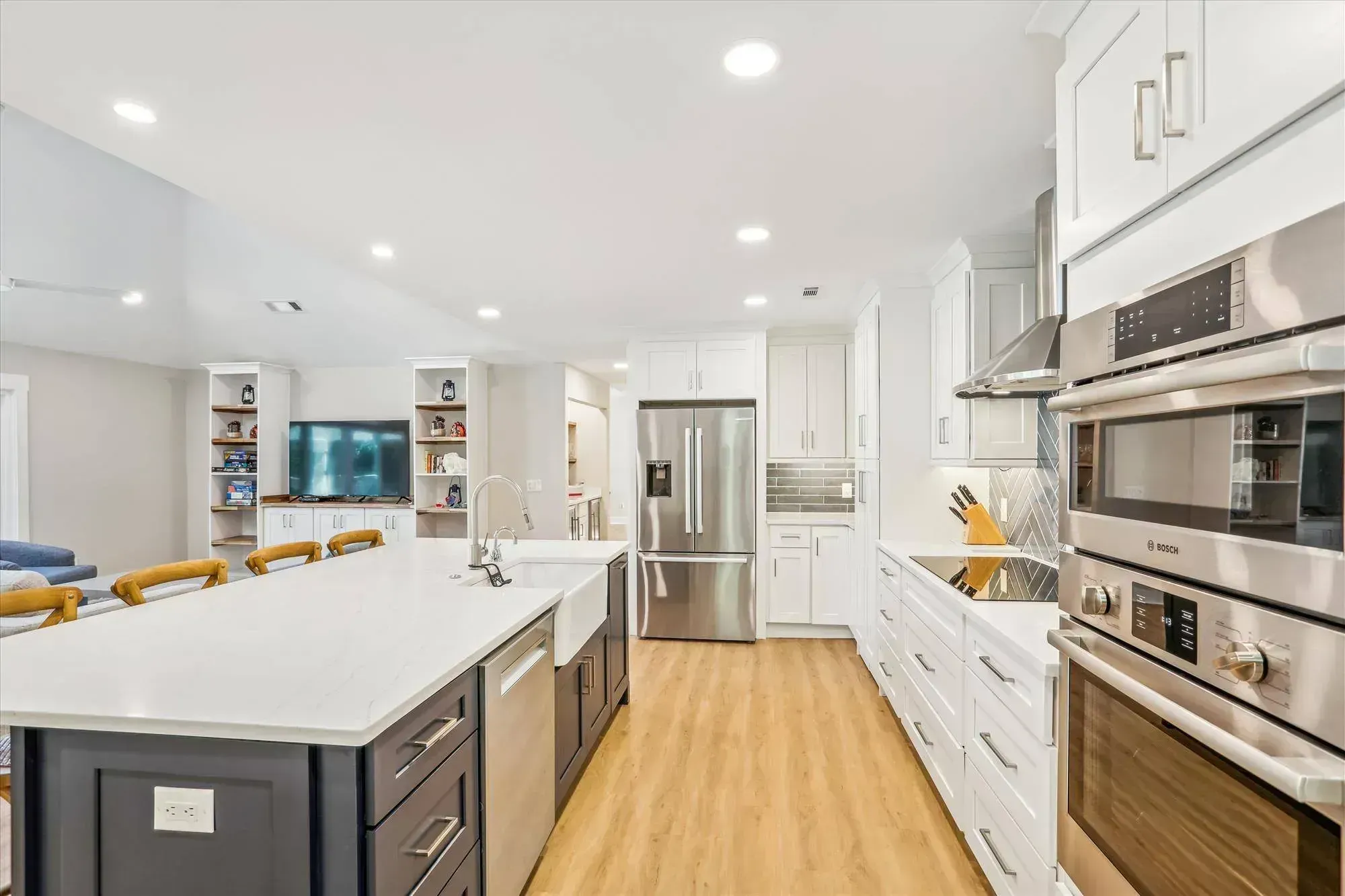 An open kitchen remodel features white shaker cabinetry, a dark two-tone island with a farmhouse sink, stainless steel appliances including double wall ovens, and a herringbone tile backsplash above light wood flooring that extends into an adjoining living area.