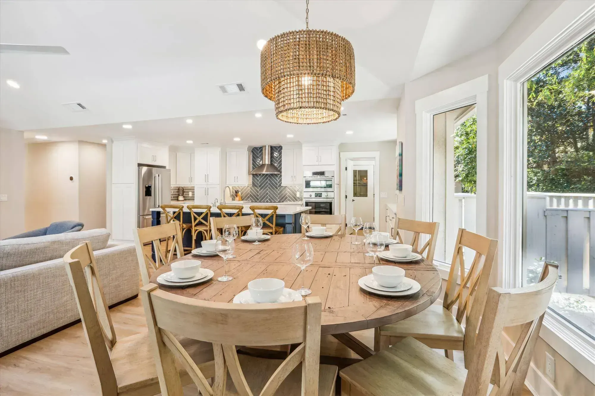 An open-concept dining area with a round wooden table set for eight sits beneath a woven pendant light, opening into a white kitchen with a navy island and herringbone tile backsplash.
