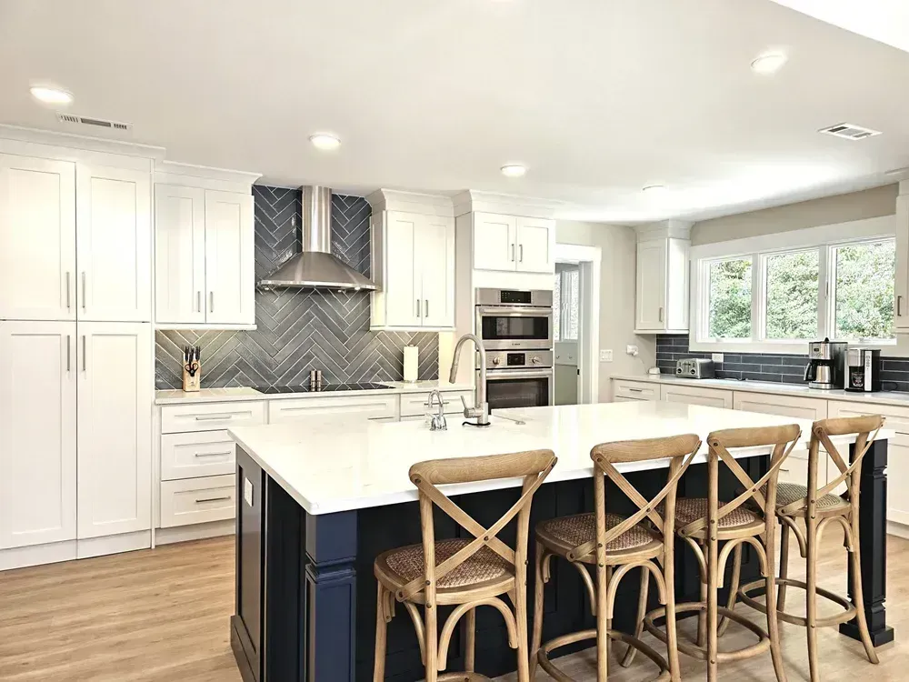 A remodeled kitchen features white shaker cabinets, a herringbone gray tile backsplash behind the stove, and a navy-blue island topped with white countertop and surrounded by four woven wood barstools.