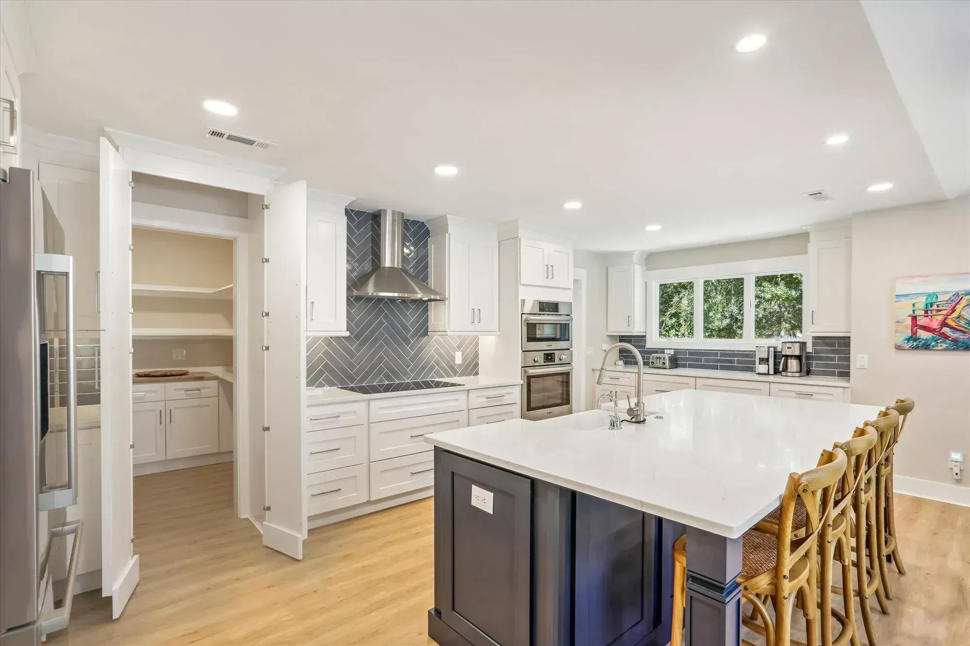 A remodeled kitchen features white shaker cabinets, a herringbone tile backsplash behind the stainless range hood, and a dark blue island with white quartz countertop and wooden bar stools, with a walk-in pantry visible to the left.