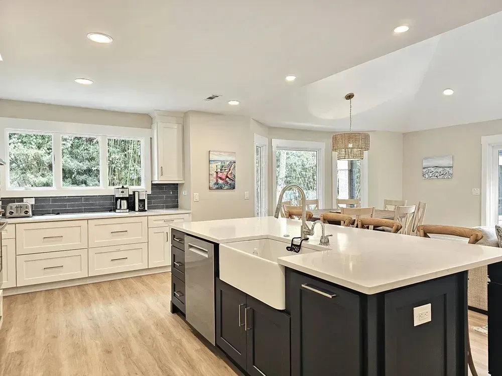 An open kitchen features white perimeter cabinetry, a dark navy blue island with a farmhouse sink, and a white quartz countertop, with a dining area and beaded pendant light visible in the background.