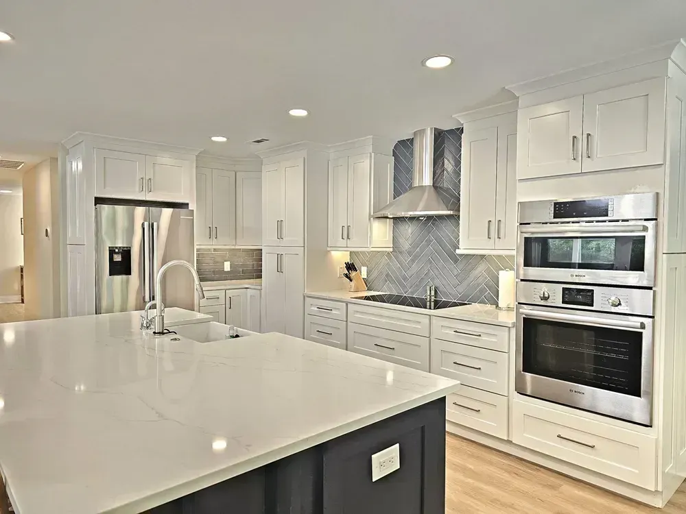 A remodeled kitchen features white shaker cabinets, a large quartz-topped island with a navy base, stainless steel double wall ovens, and a gray herringbone tile backsplash behind the cooktop.