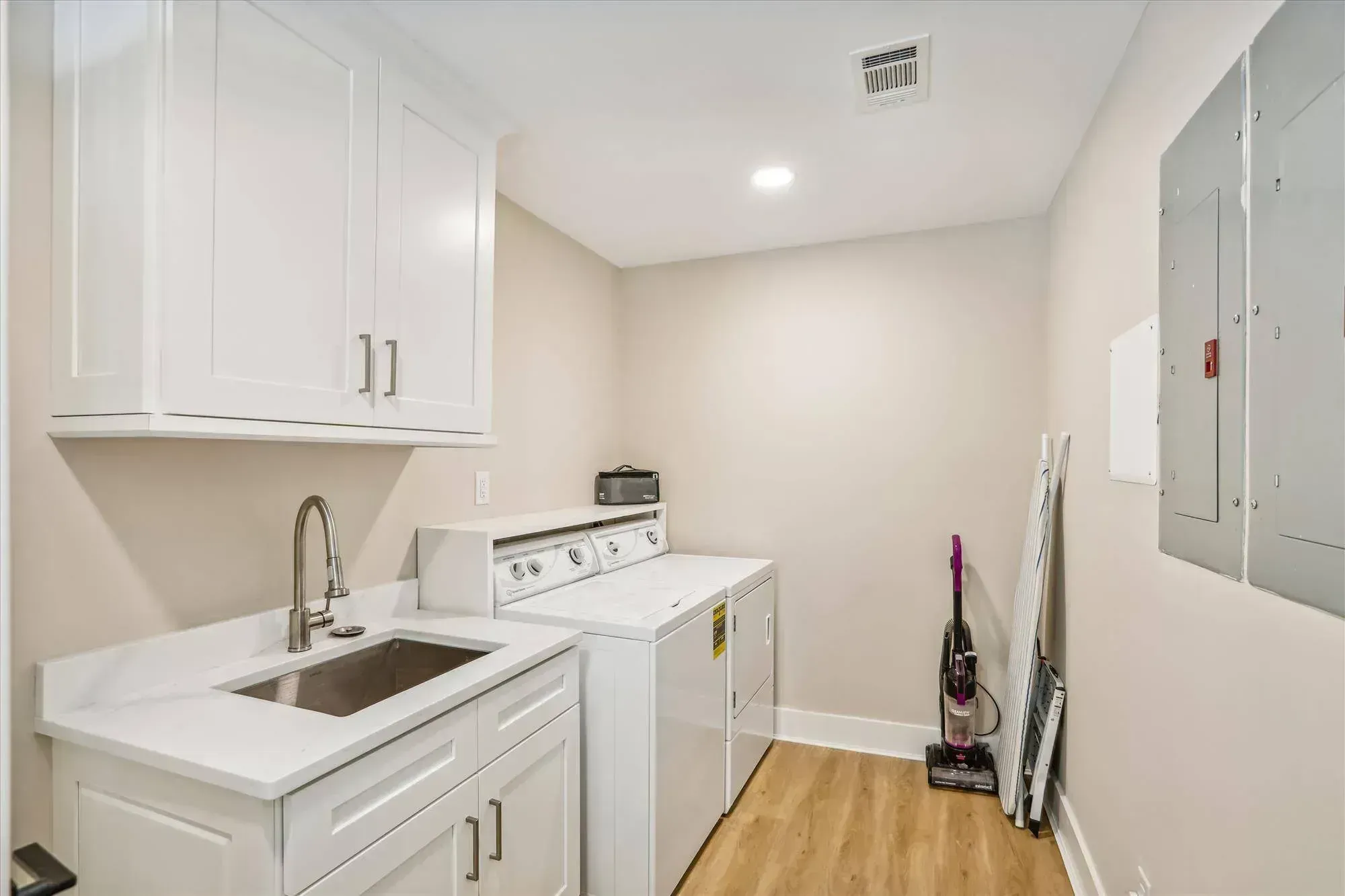A remodeled laundry room features white shaker cabinets, a stainless utility sink with marble-look countertop, a side-by-side washer and dryer, and light wood-look flooring.