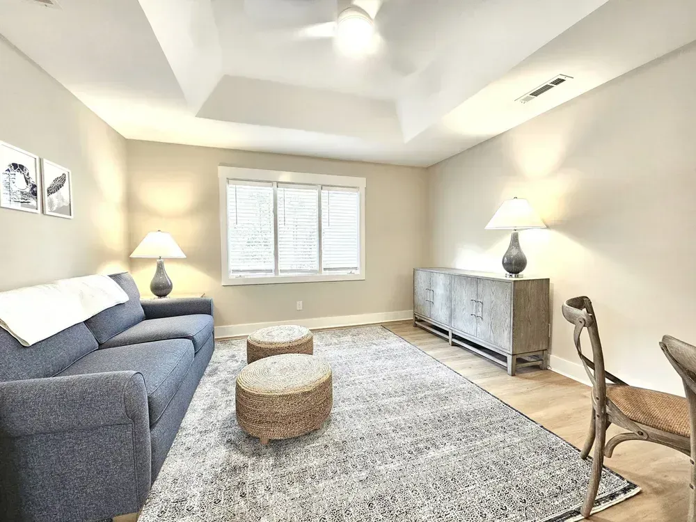 A living room with a tray ceiling features a gray sofa, woven ottomans on a patterned area rug, and a wooden sideboard flanked by lamps beneath a large window.