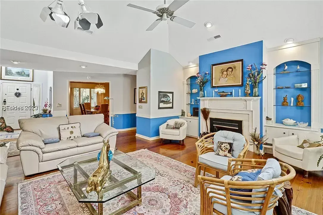 A living room with hardwood floors features a white mantled fireplace set against a blue accent wall with built-in display shelving, surrounded by upholstered seating and a glass coffee table.