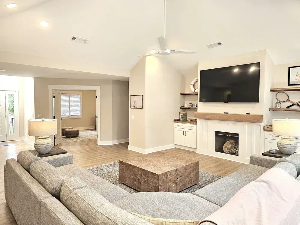 A vaulted living room features a gray sectional sofa, a wood block coffee table, and a white built-in cabinet with floating shelves flanking a wall-mounted television above a fireplace.