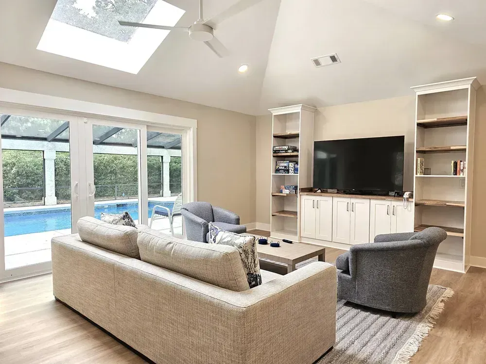 A vaulted living room with a skylight and ceiling fan features a built-in white entertainment center flanked by open shelving, a sectional sofa and gray armchairs, with sliding glass doors opening to a screened pool lanai.