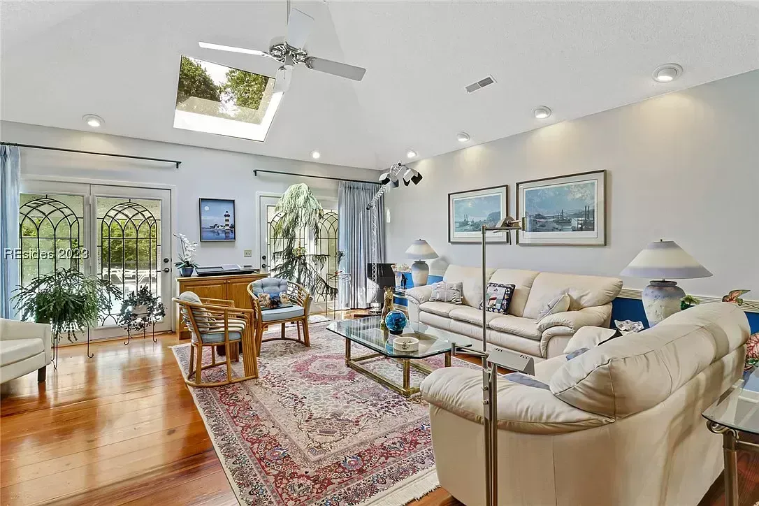 A vaulted living room with a skylight and ceiling fan features hardwood floors, a patterned area rug, cream sofas, and glass-paned French doors leading to a patio.
