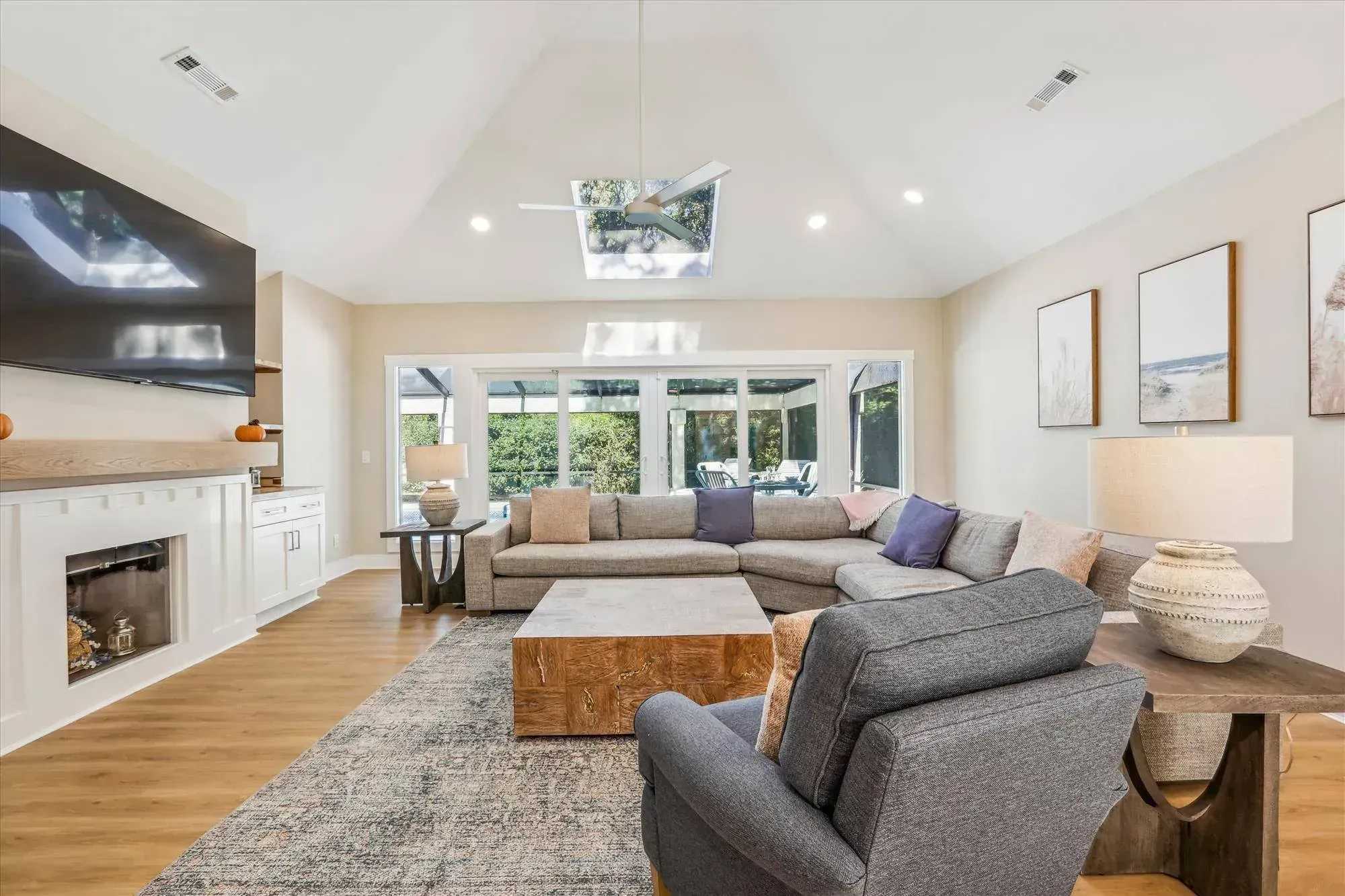 A vaulted living room with a skylight and ceiling fan features a gray sectional sofa, wood coffee table, and a built-in fireplace beneath a mounted television, with sliding glass doors opening to a screened porch beyond.
