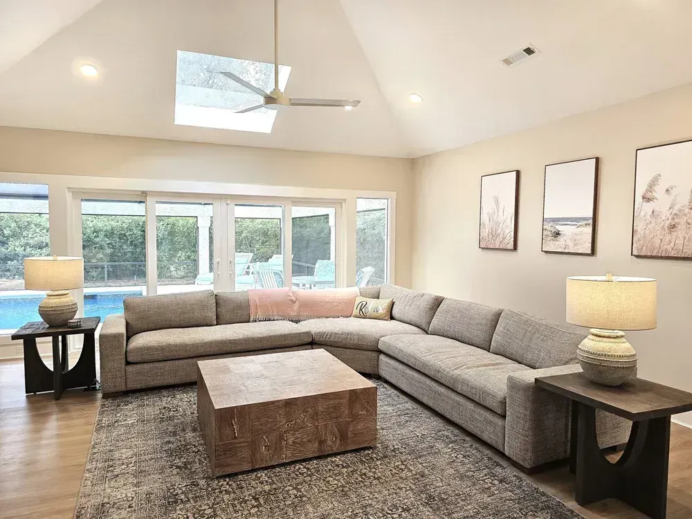 A living room with a vaulted ceiling and skylight features a gray sectional sofa, a wooden coffee table, and a wall of glass doors that overlook a pool patio.