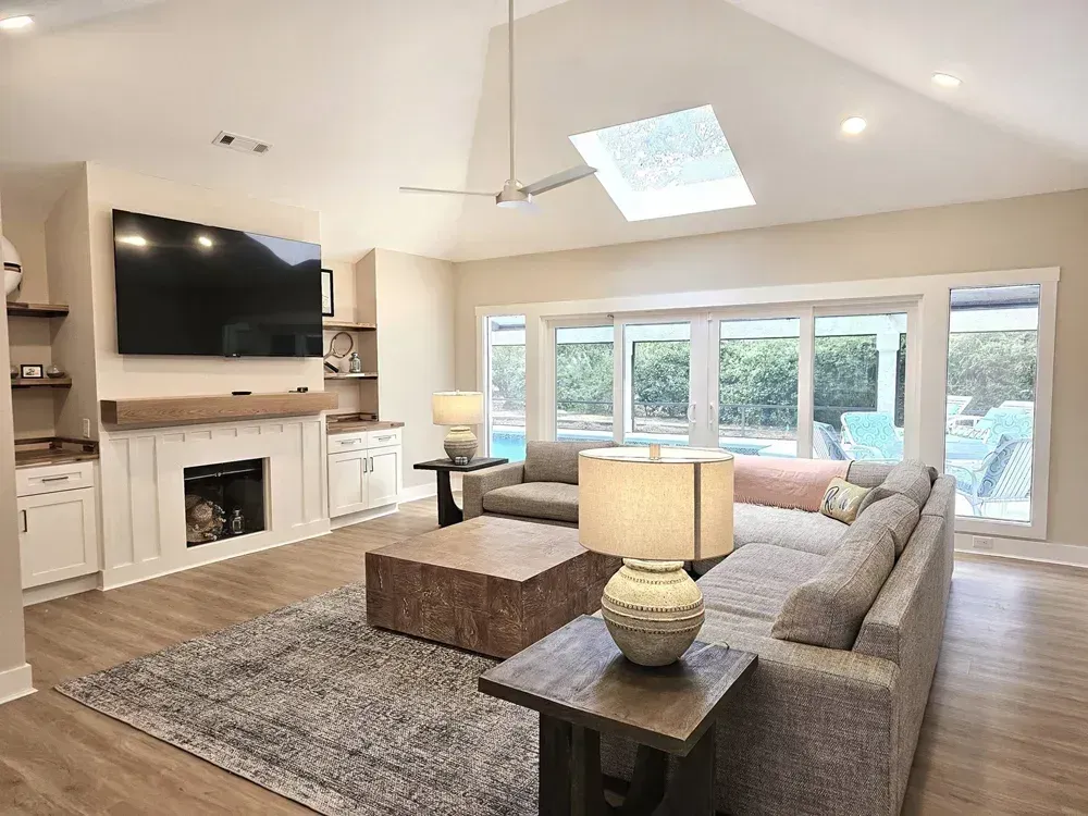 A vaulted living room with a skylight and ceiling fan features a sectional sofa, a built-in fireplace with white cabinetry, and large windows overlooking a pool.