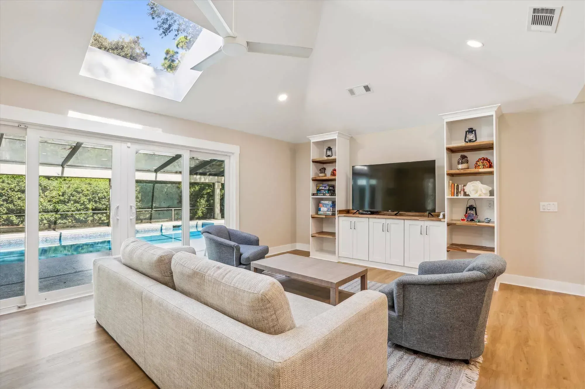 A vaulted living room with a skylight and ceiling fan features a beige sofa and gray armchairs facing a built-in white entertainment center, with sliding glass doors opening to a screened pool lanai.