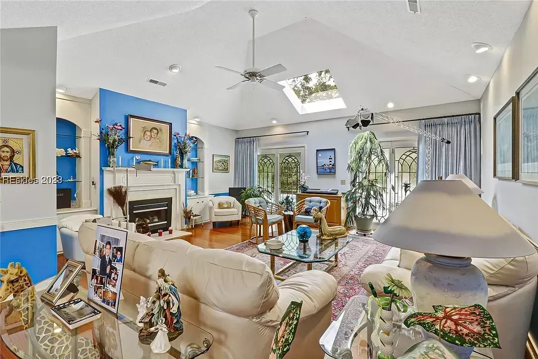 A vaulted living room with a skylight and ceiling fan features a blue accent wall, a white fireplace mantel, cream sofas, a glass coffee table, and arched windows leading to a sunroom seating area.