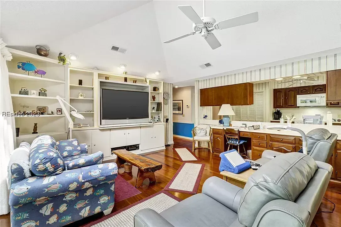 A living room with a vaulted ceiling and ceiling fan features a built-in white entertainment center with a large television, fish-patterned upholstered chairs, gray leather sofas, and a wood plank coffee table on a hardwood floor, opening into a kitchen and desk area in the background.