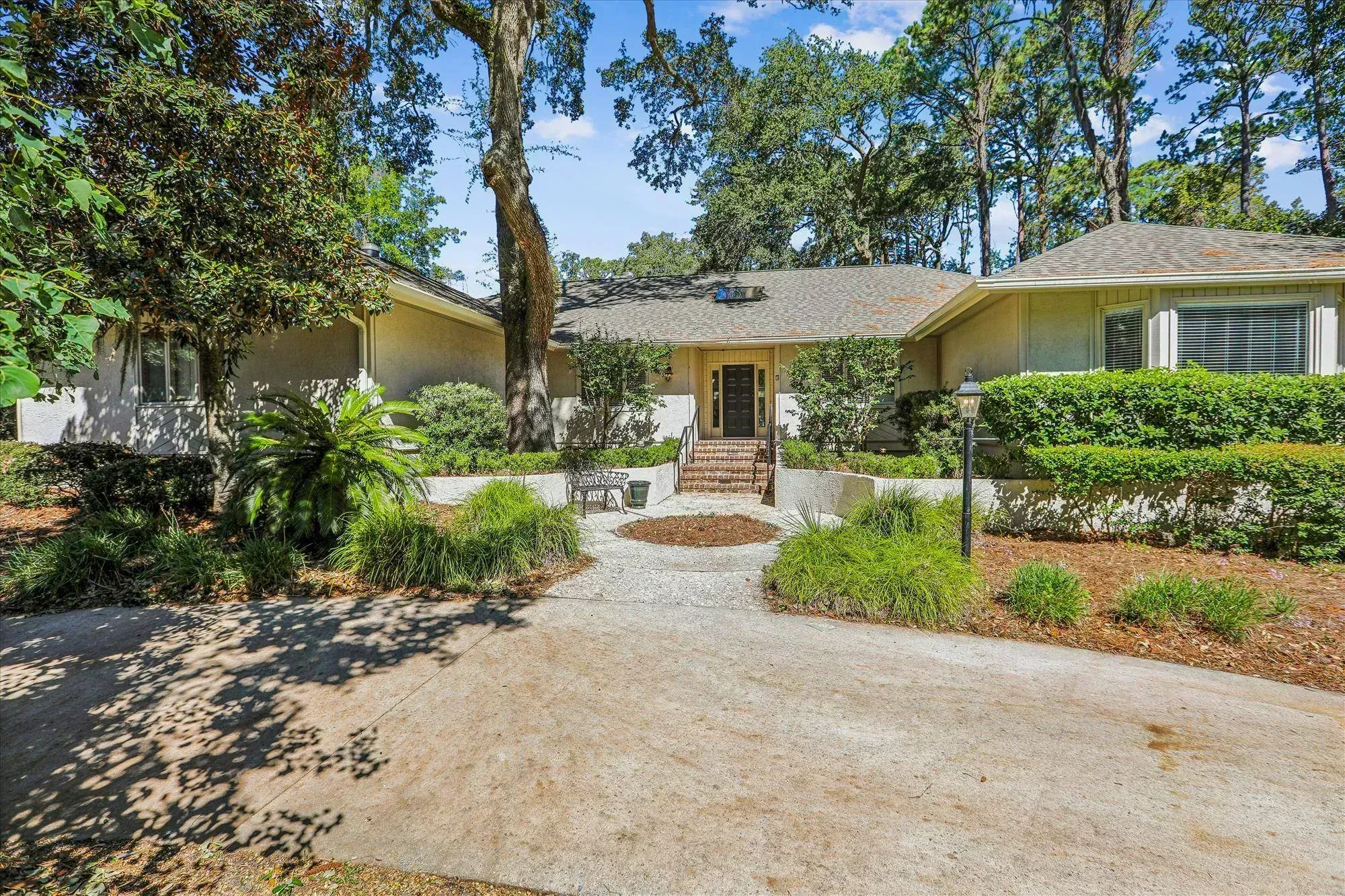 A single-story stucco home with a shingled roof sits beneath large oak and pine trees, its brick steps leading up to a dark front door flanked by mature landscaping.