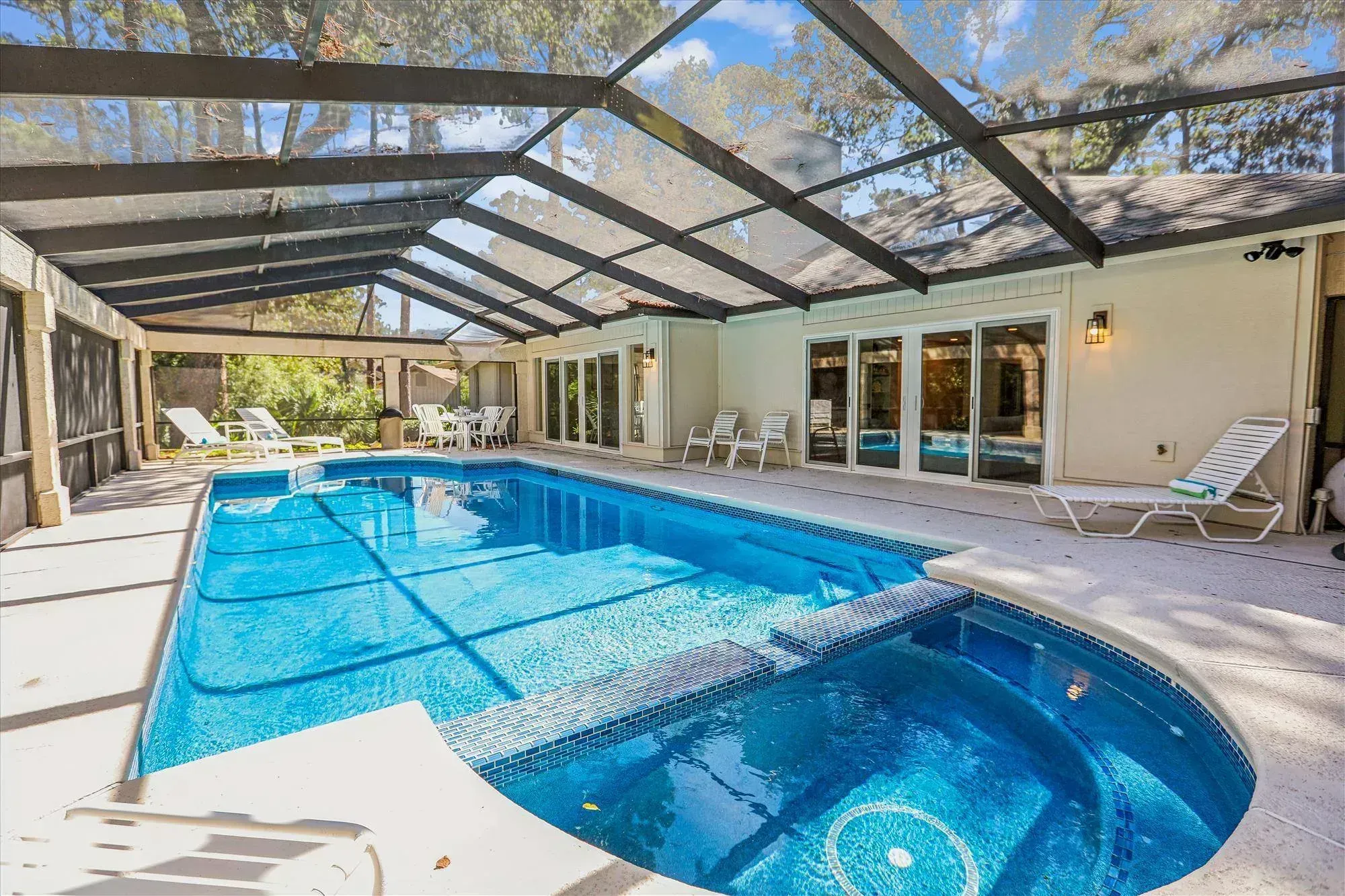 A screened-in pool enclosure with a rectangular pool and adjoining round spa surrounded by a concrete patio, lounge chairs, and a house with multiple sliding glass doors visible under a canopy of trees.