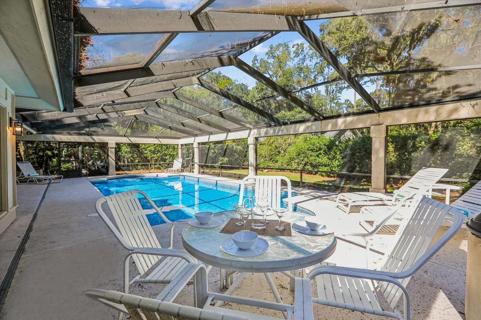 A screened pool enclosure with a rectangular swimming pool surrounded by white metal chaise lounges and a round glass-top dining table set with dishware, framed by wooded views.