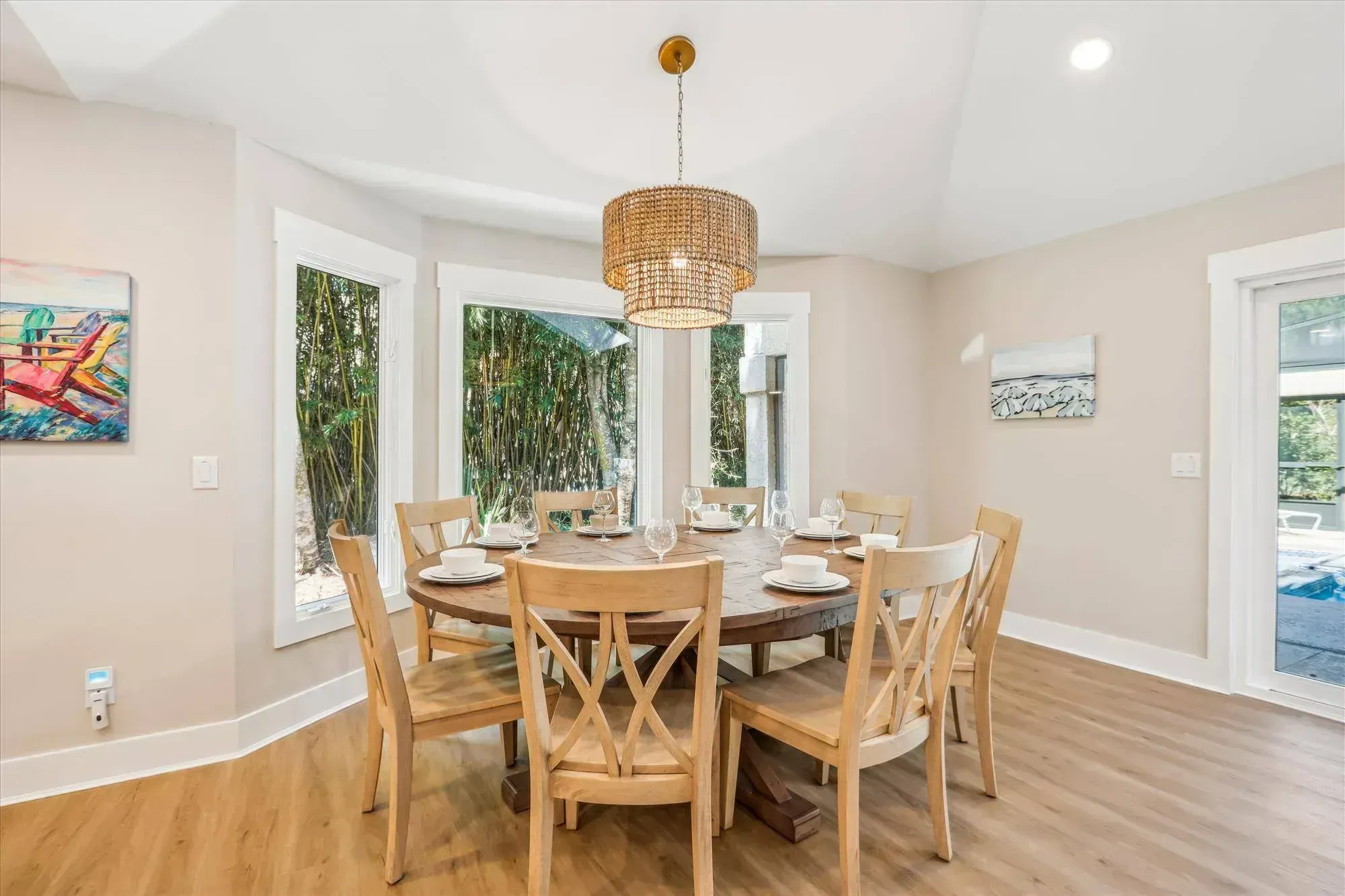 A round wooden dining table set with dishware and stemware is surrounded by eight light wood chairs beneath a beaded pendant chandelier, with bay windows overlooking greenery and a glass door leading to a pool area.
