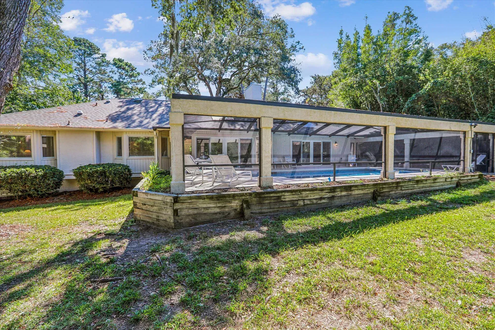A single-story ranch home has a long screened pool enclosure with lounge chairs and a rectangular pool, surrounded by a lawn and tall trees.