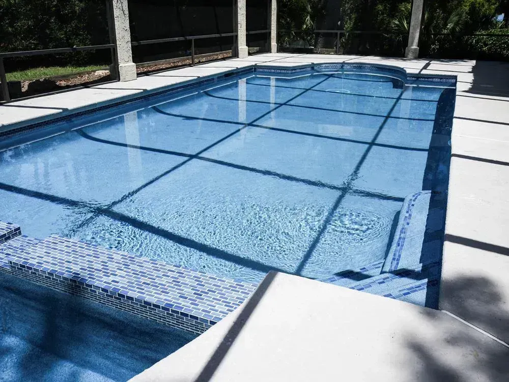 A screened outdoor pool with blue mosaic tile trim reflects a shaded lattice pattern of sunlight across the clear water and surrounding concrete deck.