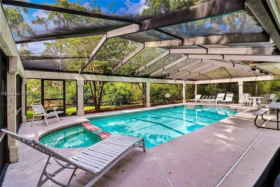 A screened outdoor enclosure shelters a rectangular pool and adjoining round spa, surrounded by lounge chairs and lush greenery visible through the mesh walls.