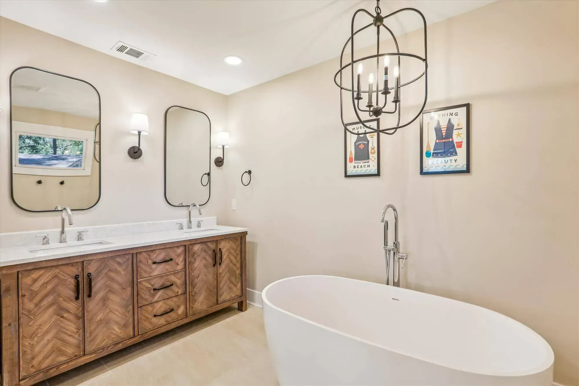 A remodeled primary bathroom features a herringbone-patterned wood double vanity with white marble countertops, two round-cornered mirrors, a freestanding oval soaking tub with floor-mounted faucet, and a globe-style chandelier overhead.