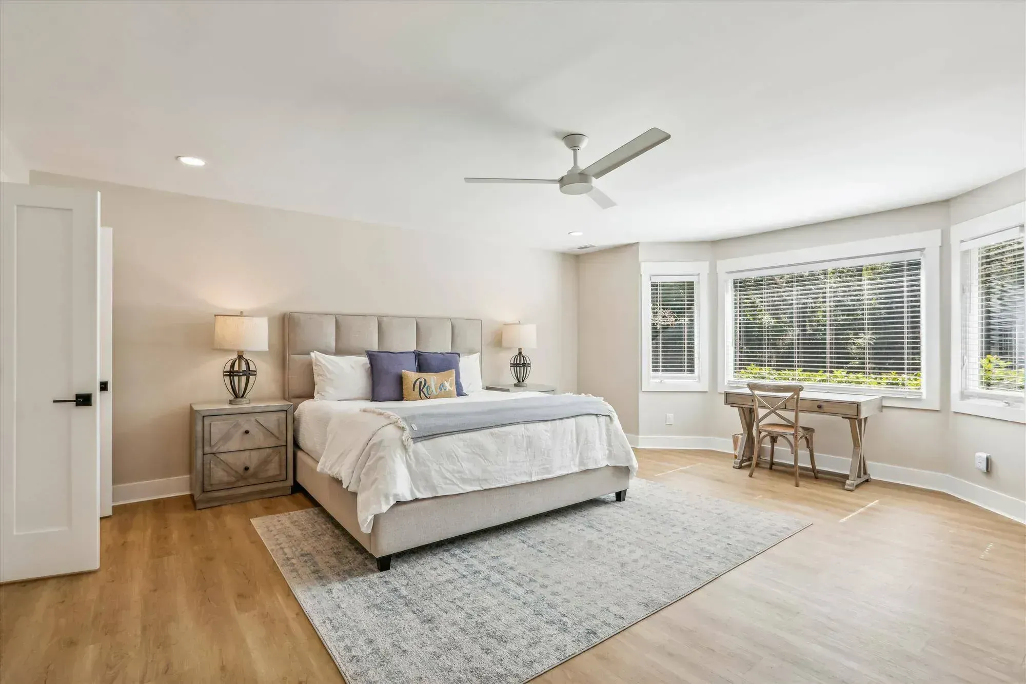 A spacious primary bedroom features a upholstered king bed with blue and white pillows, wood-look flooring, a ceiling fan, and a bay window seating area with a small desk overlooking greenery.