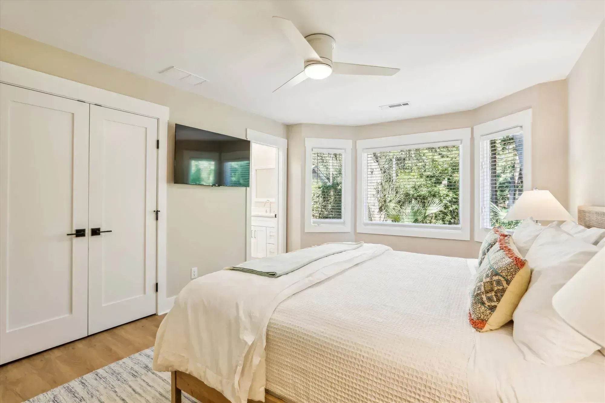 A bright primary bedroom features a bay window with wooded views, a wall-mounted television, double closet doors, and a bed dressed in white linens with an ensuite bathroom visible through an open doorway.