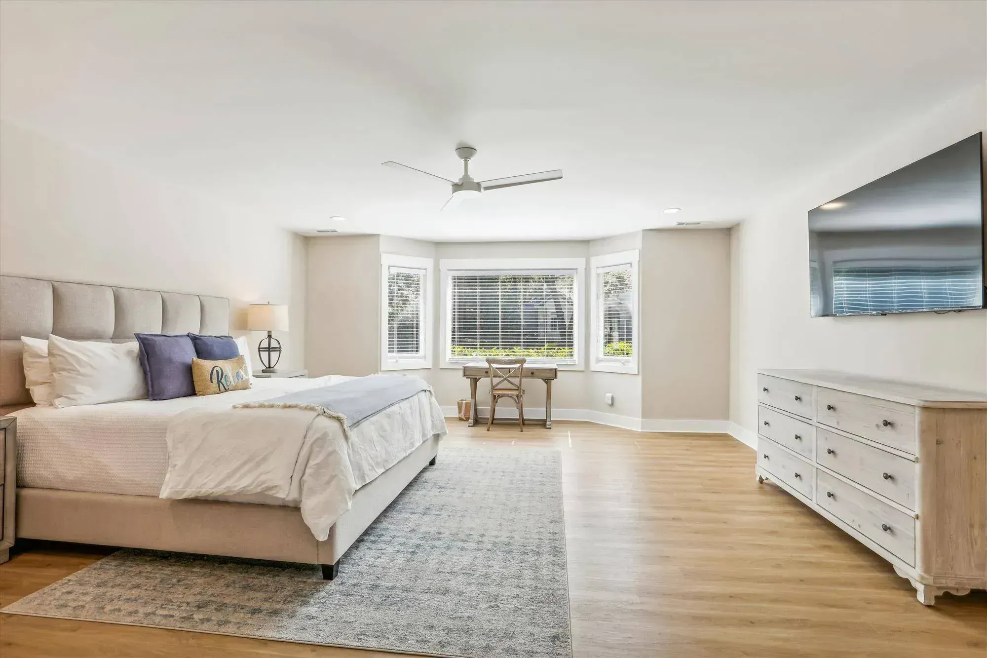 A spacious primary bedroom features an upholstered king bed with blue and white pillows, a bay window with a small desk, a wall-mounted television, and a light wood dresser atop light hardwood flooring.