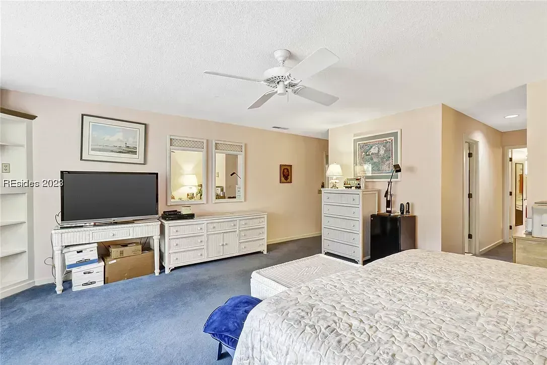 A carpeted primary bedroom features a white wicker dresser and matching nightstand with mirrors, a flat-screen television, a ceiling fan, and a bed in the foreground beneath soft neutral walls.