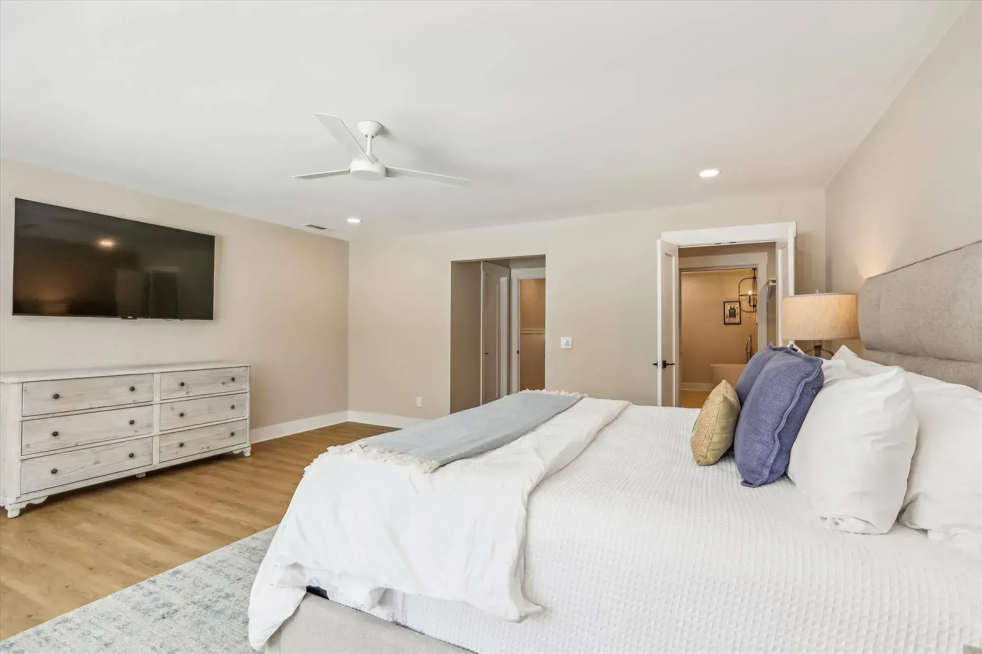 A bright primary bedroom features an upholstered bed with white and blue linens, a whitewashed dresser, a wall-mounted television, and doorways leading to a closet and an en-suite bathroom with a freestanding tub.