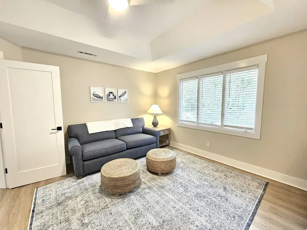 A bright sitting room addition features a tray ceiling, a gray sofa with two woven ottomans on a patterned area rug, and a large window overlooking greenery.
