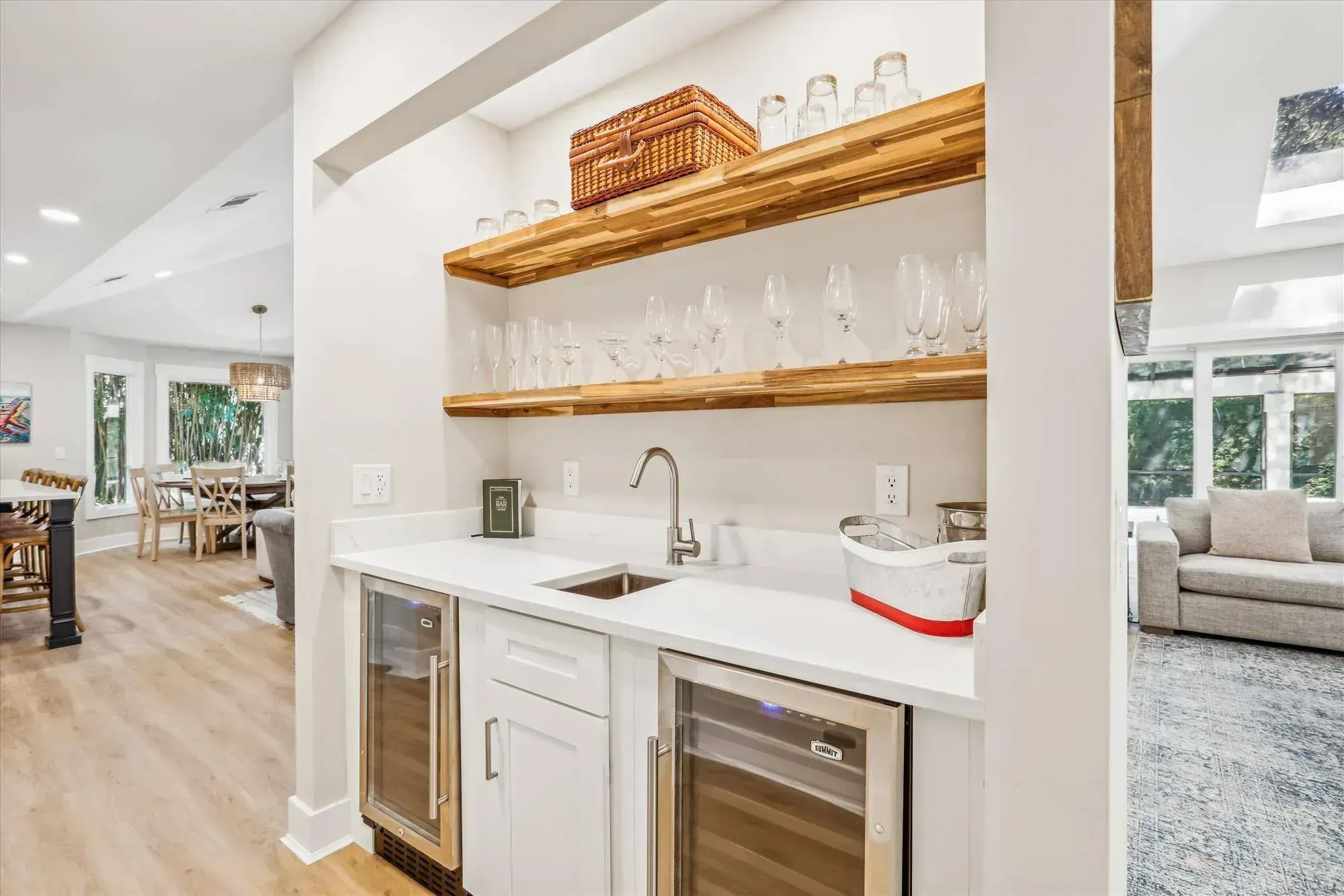 A built-in wet bar features white cabinetry, twin glass-front beverage refrigerators, a stainless bar sink, and floating wood shelves lined with glassware, opening onto a bright living and dining area beyond.