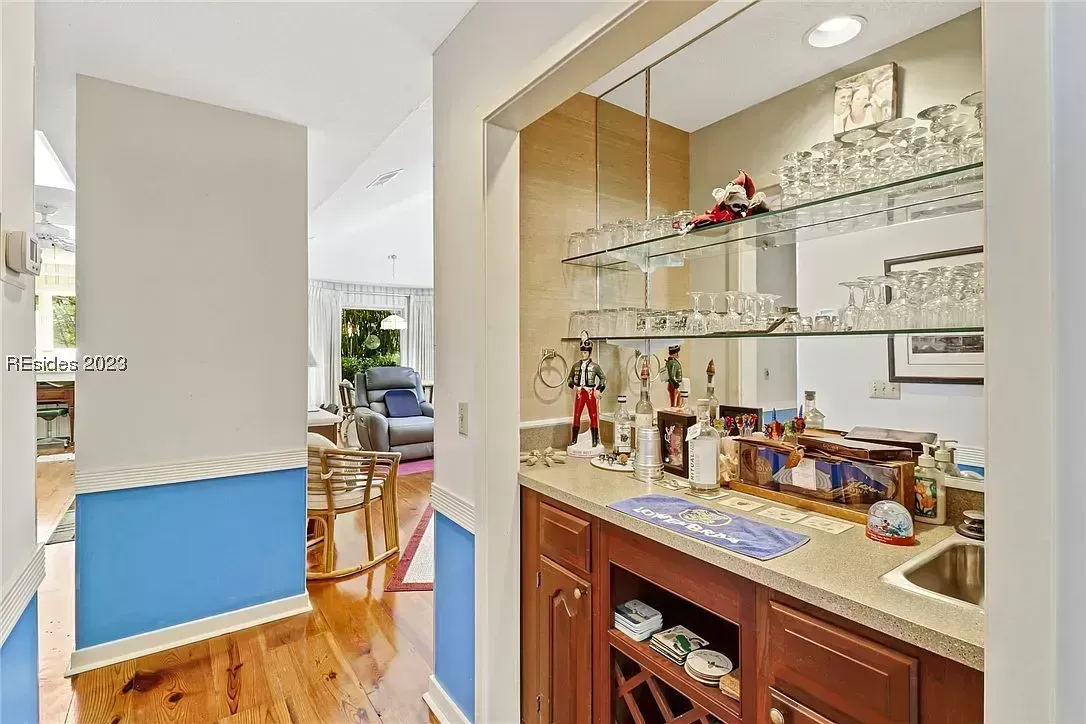 A built-in wet bar with wood cabinetry, a small sink, and mirrored glass shelves holding stemware opens onto a hallway that leads to a sunlit living room with hardwood floors.